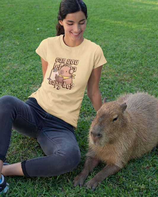 Woman sitting on grass with a capybara wearing a t-shirt with a cartoon design.